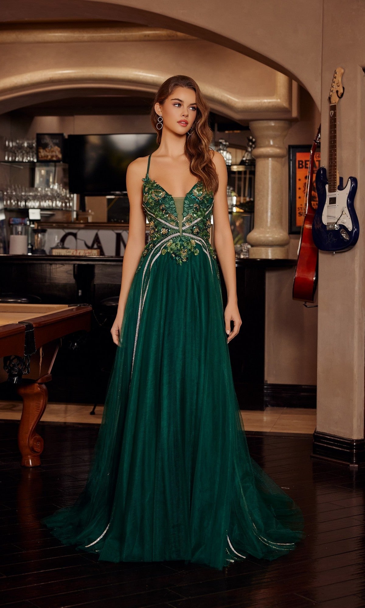 A woman in the Nox Anabel G1795 long emerald prom dress with floral embroidery and an open back stands in a room decorated with musical instruments and a pool table.